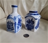 Detailed close up view of two Russian porcelain Gzhel bottles in white and blue featuring floral and bird designs with a coin for scale resting on a white surface