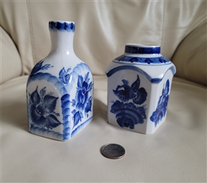 Detailed close up view of two Russian porcelain Gzhel bottles in white and blue featuring floral and bird designs with a coin for scale resting on a white surface