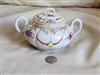 The front view shows an antique bulbous porcelain sugar bowl with two handles and a lid featuring hand painted pink rose swags and intricate gold tone scrolls. An angled view highlights the embossed texture of the porcelain and the ornate gilded handles t