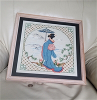 Framed vintage Japanese cross stitch art featuring a Geisha with a blue kimono and umbrella in a mountain landscape