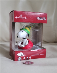 Hallmark PEANUTS Snoopy molded plastic Christmas ornament wearing green hat and red scarf seated on red sled with box