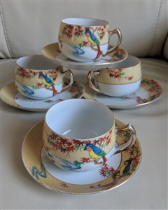 A group of four delicate Japanese teacups and saucers with colorful parrot designs, showing the translucent nature of the porcelain and gold toned handles